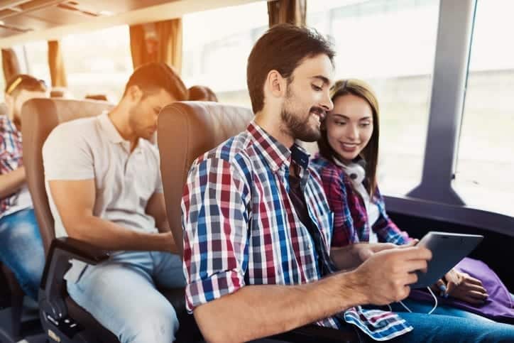 Charter Bus to Pennsylvania - Leprechaun Lines - The guy and the girl are on the bus. The guy is holding a tablet. They are looking at something. The young guy's girlfriend is smiling. With them, the bus goes other tourists who also went on a trip.